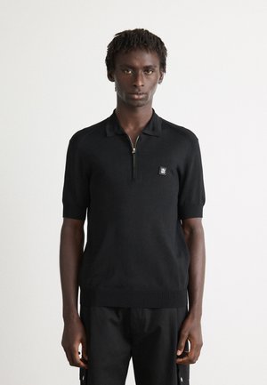 Hombre vistiendo una camiseta polo de manga corta y cremallera negra con un pequeño logo de Hugo en el pecho, de pie contra un fondo blanco liso.