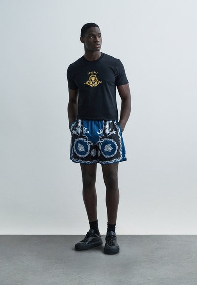 Tricou negru cu logo auriu, asortat cu pantaloni scurți albaștri cu model și detalii albe, și adidași negri. Material neted și croială lejeră.