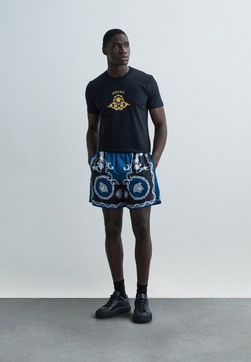 Sort T-shirt med gyldent logo, kombineret med blå mønstrede shorts med hvide detaljer og sorte sneakers. Glat stof og afslappet pasform.