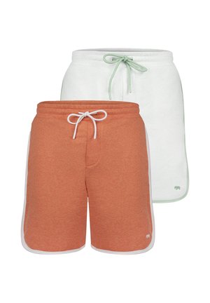 Uomo che indossa pantaloncini arancioni con coulisse e bordo bianco, maglietta bianca e scarpe da ginnastica bianche, in piedi davanti a uno sfondo bianco.