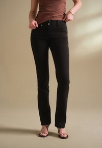 Frau trägt eng anliegende schwarze Jeans und schwarze Riemchenabsätze, steht vor einem schlichten beigen Hintergrund mit den Händen nahe am Bund.