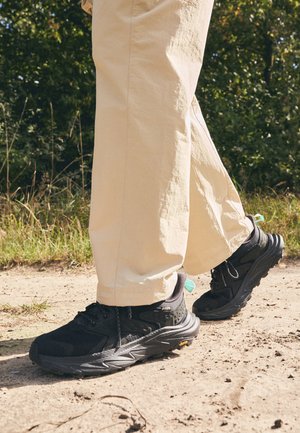 Melni pārgājienu apavi ar teksturētu augšdaļu, šņorēm un bieza zole ar Vibram logotipu. Valkāti ar gaišiem biksēm.