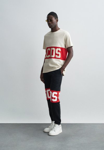 T-shirt in cotone beige con fascia orizzontale rossa che presenta "CDS" in lettere bianche; abbinato a pantaloni neri con fascia rossa simile e sneaker bianchi.