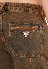 Bruine denim hoge taillebroek met een rits achterzak en een driehoekig 'GUESS' logo patch. Getextureerde stof met een subtiel patroon.