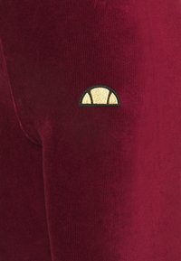 Burgundy corduroy leggings yang menampilkan permukaan tekstur dan logo jahit hitam dan emas di area paha atas.