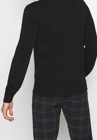 Suéter negro de punto con mangas largas, con un dobladillo y puños acanalados. Combinado con pantalones de cuadros en tonos verde oscuro y azul marino.