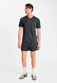 Ciemnoszary sportowy t-shirt z dekoltem w serek, krótkimi rękawami i subtelnym logo. Do tego czarne siateczkowe spodenki oraz jasne buty sportowe.