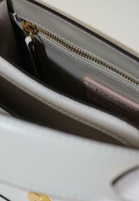 Indersiden af en beige læderhåndtaske med en guldlås-lomme og en lyserød label med mærke- og materialedetaljer.