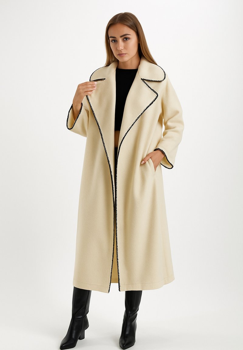 Donna che indossa un lungo cappotto crema con rifiniture nere, top nero corto e stivali neri fino al ginocchio, in piedi contro uno sfondo bianco semplice.