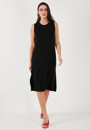 Black sleeveless dress, knee-length, made of smooth fabric. Side slits enhance movement. Paired with red heeled sandals.