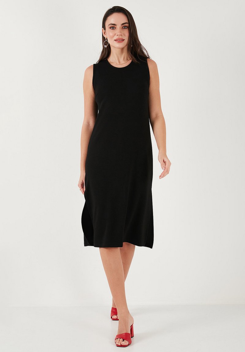 Black sleeveless dress, knee-length, made of smooth fabric. Side slits enhance movement. Paired with red heeled sandals.