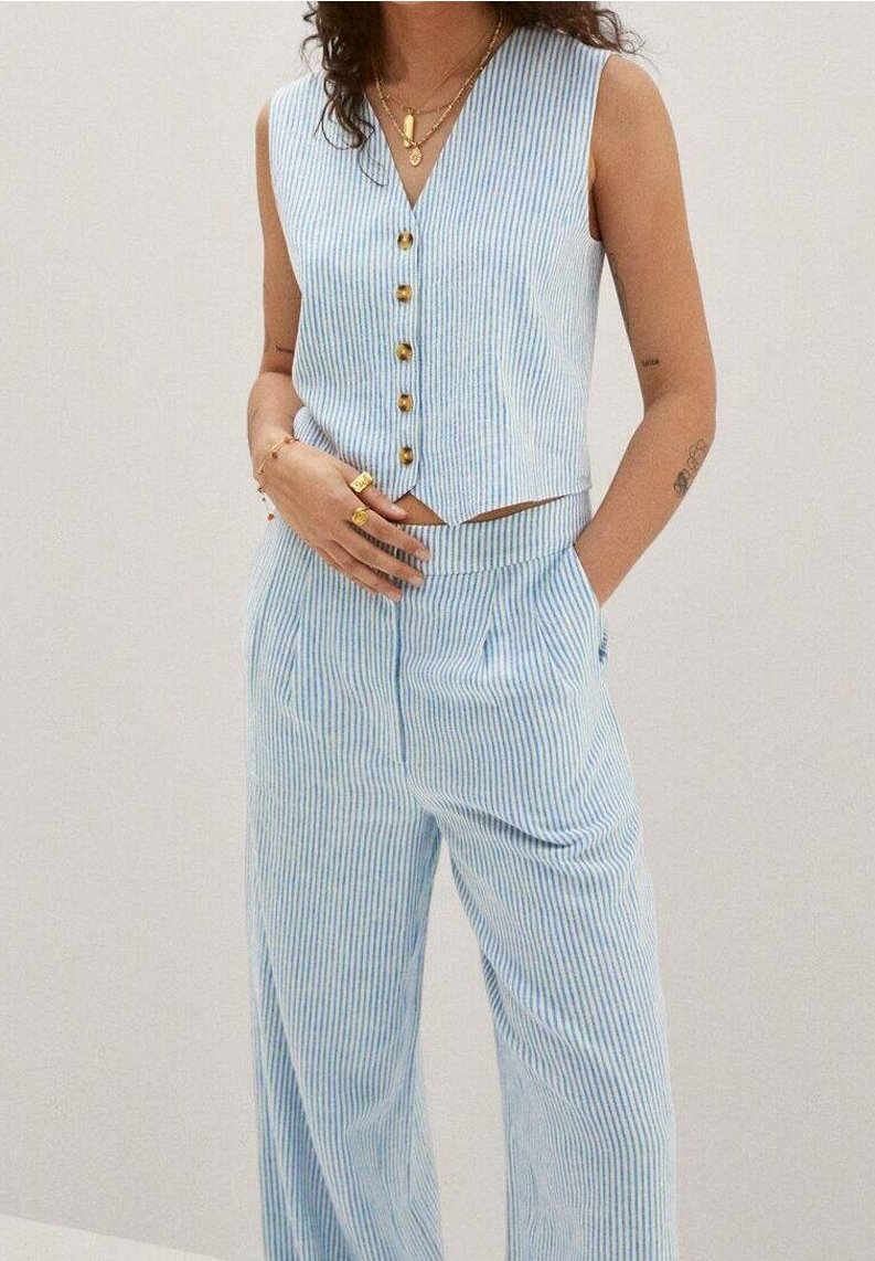 Femme portant un haut sans manches boutonné à rayures verticales bleu clair et blanc, et un pantalon large assorti avec des bagues et colliers dorés.