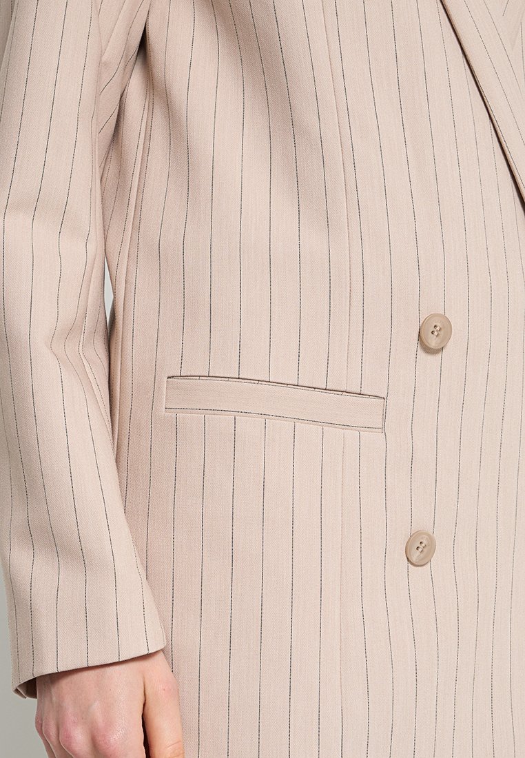 Blazer beige à rayures avec poche passepoilée et deux boutons, porté par une personne avec une main visible près du coin inférieur gauche.
