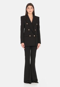 Blazer nero doppiopetto con bottoni dorati, vestibilità sartoriale e spalle definite, abbinato a pantaloni neri svasati e tacchi a punta.