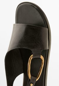 Schwarze Ledersandale mit breitem Riemen, strukturiertem Obermaterial und goldener ovaler Akzent an der Vorderseite. Flache Sohle mit minimalistischem Design.