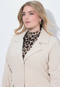Una donna bionda con i capelli sciolti indossa orecchini a cerchio d'oro, un dolcevita con stampa leopardata e un blazer in corduroy beige, mentre guarda leggermente di lato.