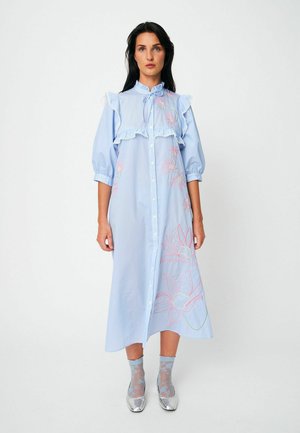 Ženska v svetlo modri midi obleki z rožnim rožnim vezenjem v roza barvi, s cofki, napihnjenimi rokavi, prozornimi nogavicami z rožami in srebrnimi čevlji, stoji mirno.