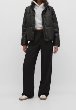 Veste matelassée en similicuir noir avec boutons-pression, sweat à capuche gris raccourci en dessous, associée à un pantalon noir ample et des baskets blanches.