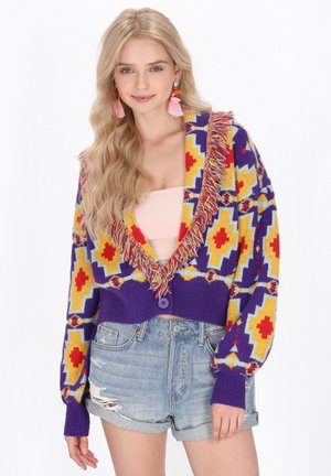 Cardigan tricotat colorat cu o bază violetă, modele geometrice galbene, roșii și albastre, având o margine din franjuri și un închizător cu un singur buton.