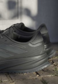 Paar dunkelgraue Sportschuhe, die auf einer gepflasterten Oberfläche mit Sonnenlicht und Schatten im Hintergrund ruhen.