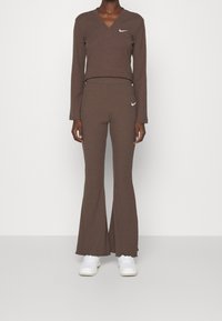 Haut long à manches en maille côtelée marron avec un décolleté en V et un pantalon évasé assorti, tous deux ornés d'un logo Nike blanc sur la poitrine et la hanche. Baskets blanches.