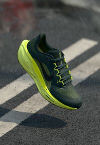 Dark green and neon yellow Nike running shoe mid-air above asphalt with white road markings, visible laces and textured sole.