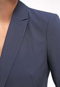 Tummansininen bleiseri, jossa on istuva muotoilu, Notched-kaulus ja teksturoitu kangas. Siistit linjat ja minimalistiset yksityiskohdat ovat näkyvissä.