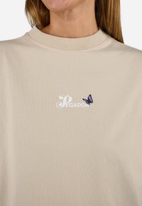 T-shirt beige con collo rotondo, tessuto morbido, logo 'Pegador' in bianco e un accento di ricamo di una farfalla viola.