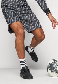 Grå mønstrede shorts med et geometrisk design, parret med sorte sportssko og hvide sokker med sorte striber, tæt på en fodbold.