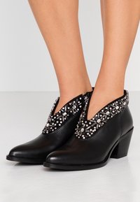 Botas de tobillo de cuero negro con un diseño en forma de V en la parte superior, adornadas con embellishments de strass plateados y claros, y un tacón moderado.