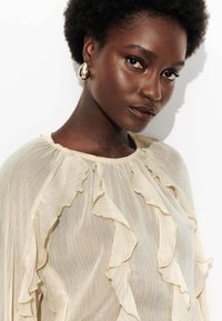 Blouse légère et transparente avec des détails froncés et un col rond dans une couleur beige doux. Dotée d'un tissu texturé et de délicats accents dorés.
