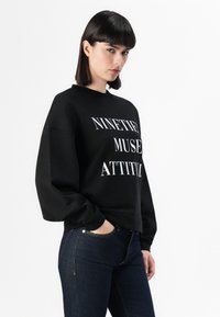 Felpa nera con la scritta "NINETIES MUSE ATTITUDE" in bianco. Polsini a coste e collo rotondo, abbinata a jeans in denim scuro.