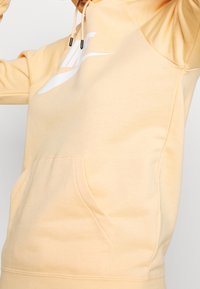 Kaptur w jasno brzoskwiniowym kolorze, wykonany z miękkiego materiału. Posiada kieszeń kangurkę oraz białe logo z przodu.