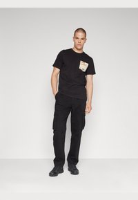 Black cotton t-shirt with a tan camo pocket, paired with black cargo pants featuring side pockets and textured fabric, worn with black sneakers.