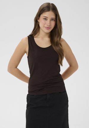 Jonge vrouw met lang bruin haar, met gouden oorringen, een zwarte mouwloze top en een zwarte rok, staand met handen achter haar rug.