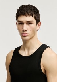 Schwarzes geripptes Tanktop mit rundem Ausschnitt und ärmellosem Design, präsentiert an einem Modell mit kurzen dunklen Haaren vor einem neutralen Hintergrund.