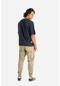 Hombre con cabello oscuro que lleva una camiseta grande y oscura, pantalones cargo beige y zapatillas navy con rayas blancas, de pie y mirando de lado.