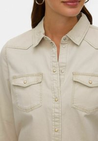 Jasnobeżowa koszula na guziki z kołnierzykiem, dwoma kieszeniami na piersi z zatrzaskami oraz subtelnymi detalami szwów.