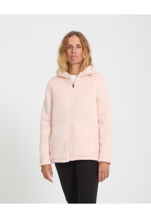 Donna con capelli biondi ondulati, indossa una felpa con cappuccio rosa chiaro con zip e pantaloni neri, in piedi davanti a uno sfondo bianco semplice.