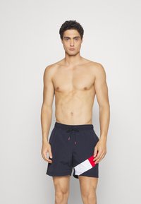 Marine badetights med elastisk midje og snøring, med et diagonalt rødt og hvitt geometrisk mønster på venstre ben. Lett materiale.