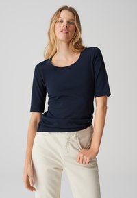Top blu navy a maniche corte con scollatura rotonda, realizzato in un tessuto liscio, abbinato a pantaloni beige chiaro e presentato su uno sfondo neutro.