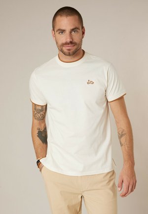 Homme aux cheveux courts et tatouages, portant un t-shirt blanc avec des bordures marron et un petit logo de scooter, un pantalon beige et des bracelets au poignet.
