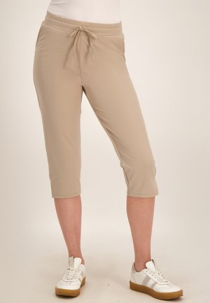 Persoon draagt beige cropped broek met een trekkoord in de taille en witte sneakers met lichtbruine zolen.