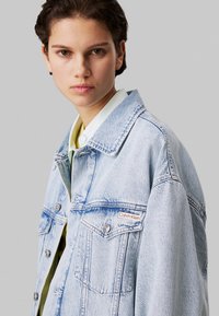 Lichtblauwe denimjacket met grote kraag, zilveren knopen, twee borstzakken en een rode Calvin Klein-label. Gladde textuur en ontspannen pasvorm.