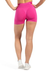 Vrouw staat met haar handen op haar heupen, gekleed in felroze strakke shorts en witte sportschoenen, tegen een witte achtergrond.