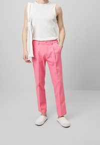 Różowe spodnie na miarę z przednimi zakładkami, zestawione z białym topem bez rękawów. Modelka ma na sobie białe buty wsuwane, stoi na tle szarego tła.