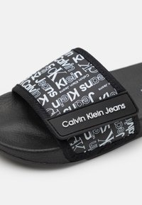 Černé sandály s texturovanou stélkou a nastavitelným páskem, které mají bílé vzory loga Calvin Klein Jeans a štítek.