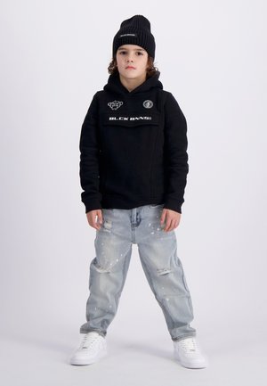Zwarte hoodie met logodetails, gecombineerd met lichtblauwe gescheurde jeans met verfspatten en witte sneakers. Een zwarte beanie maakt de look af.