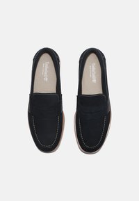 Mocasines de ante negro con un diseño cosido, punta redondeada y suela en un tono marrón claro contrastante. Presentan un panel acento elevado en la parte frontal.