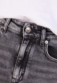 Jeans de mezclilla negra con un acabado desgastado, con un botón plateado, cierre de cremallera y un diseño clásico de cinco bolsillos. Tejido texturizado visible.